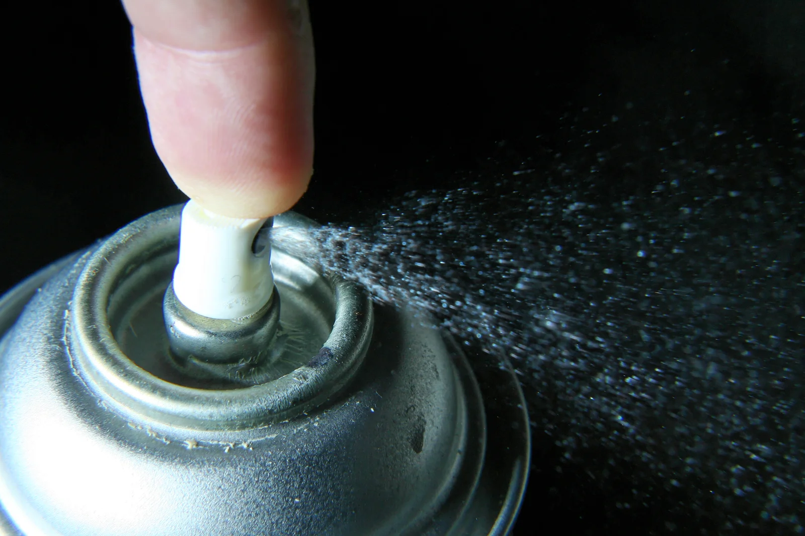 Close-up of a finger pressing the nozzle of an aerosol can, a fine mist spraying out against a black background