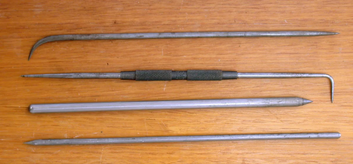Four metalworking scribers with pointed steel tips laid side by side on a plain background