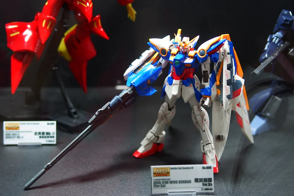 A built MG Wing Gundam Ver.Ka with a long rifle, posed in a display case beside a card reading XXXG-01W Wing Gundam Ver.Ka, No.069, 2004.03