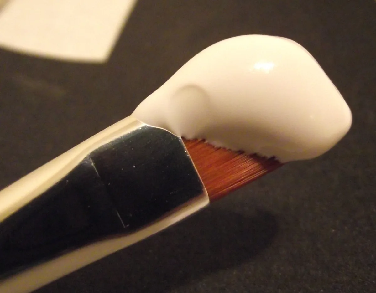 A flat brush with a thick blob of white acrylic gesso on its tip, photographed against a dark background
