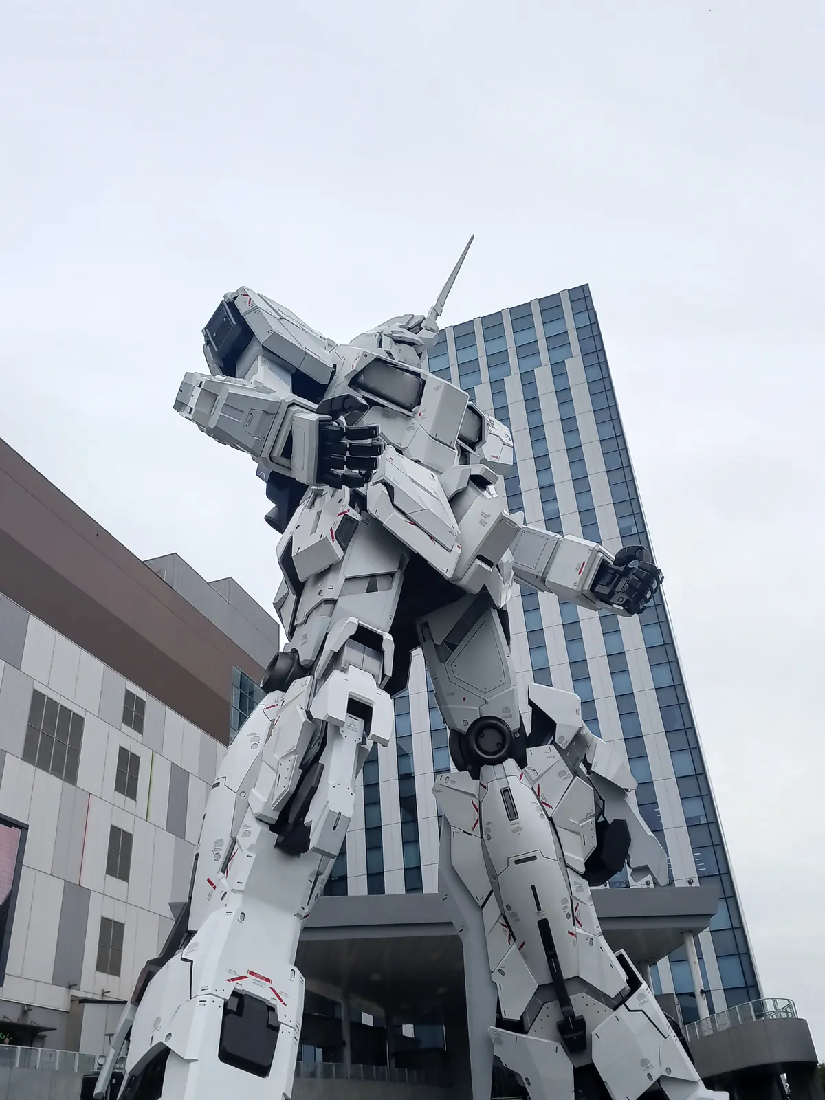 A life-sized Unicorn Gundam statue in Unicorn Mode, photographed from below in front of a glass office tower